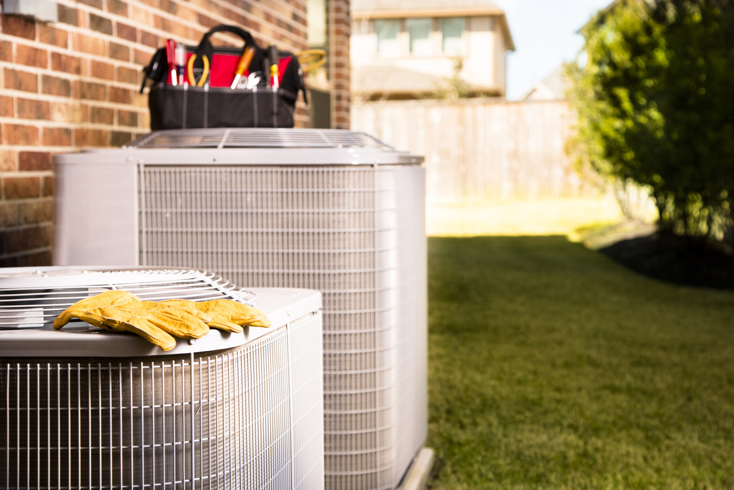 Bag of repairman's work tools, gloves on top of air conditioner units outside a brick home.  Service industry, working class.